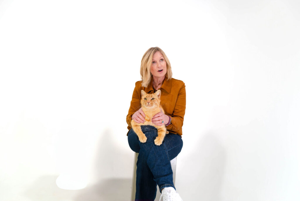 A white woman is seated with an orange cat on her lap with a white backdrop.