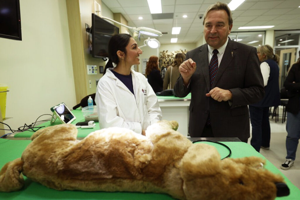 A woman of colour clad in a lab coat stands next to a white male in a suit, with a canine plushie in the foreground, used during veterinarian students' clinical skills training.