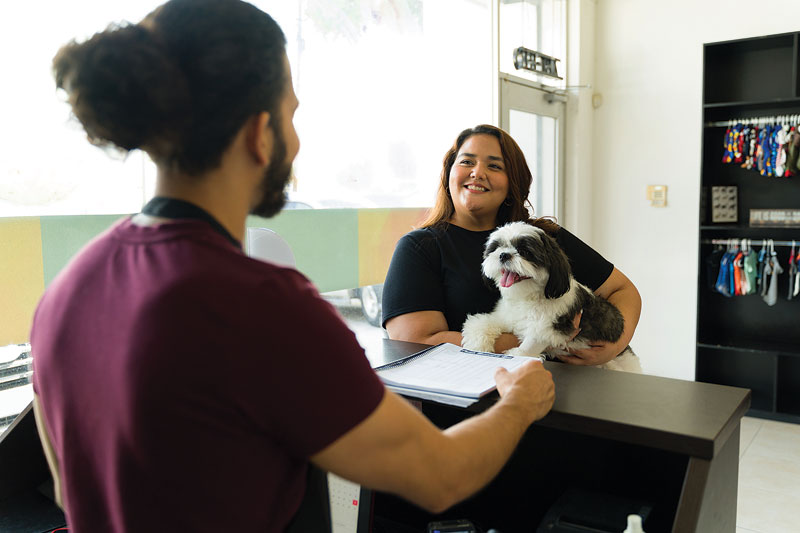 A female carrying a dog smiles to a male veterinary staff in a clinic.