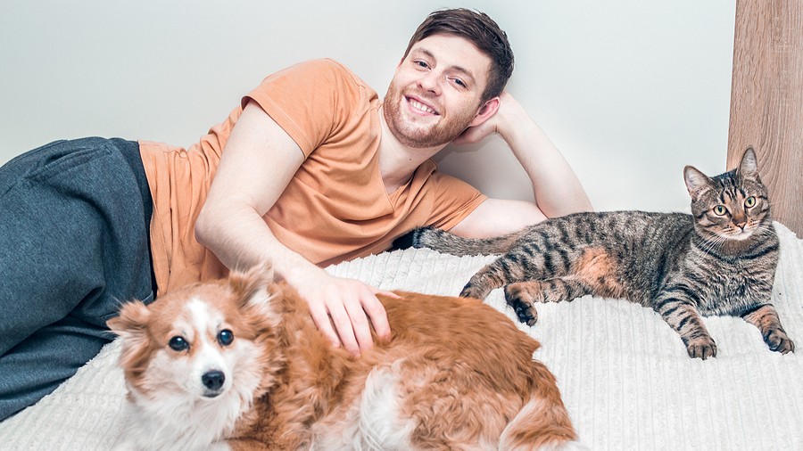 Smiling man lying on a bed with a fluffy brown-and-white dog in the foreground and a striped tabby cat beside him.